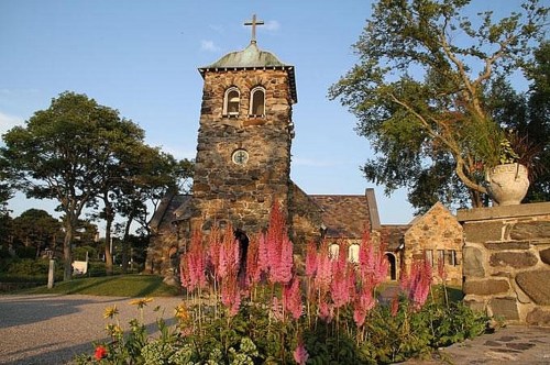 A stone church with a cross atop its tower, surrounded by colorful flowers and trees under a clear blue sky.