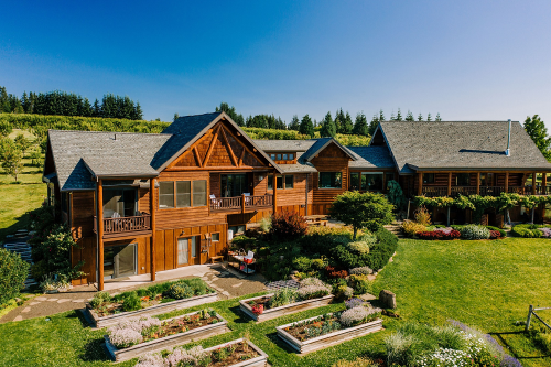 A large wooden house surrounded by gardens and greenery under a clear blue sky.