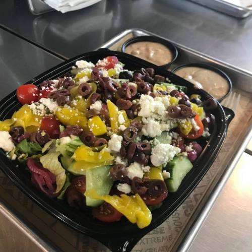 A colorful salad with mixed greens, cherry tomatoes, olives, cheese, and yellow peppers, served with two small containers of dressing.