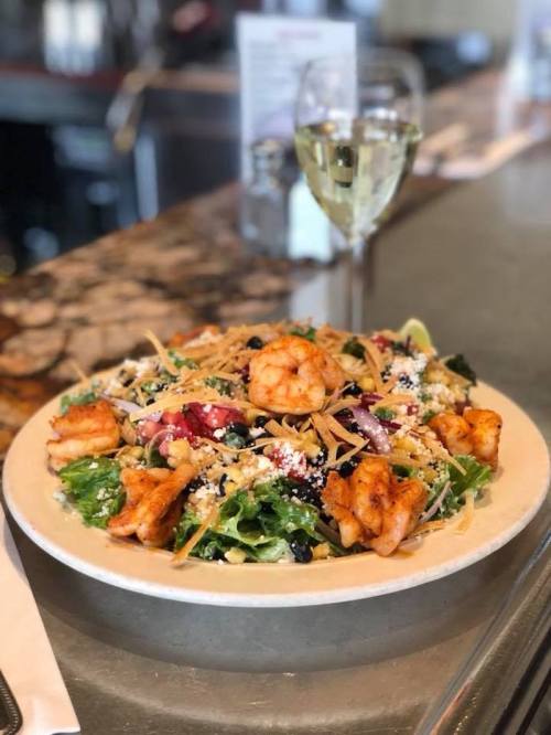 A vibrant salad topped with shrimp, greens, and crispy toppings, served with a glass of white wine in the background.