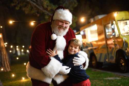 A smiling child hugs Santa Claus in a festive outdoor setting with string lights and a food truck in the background.