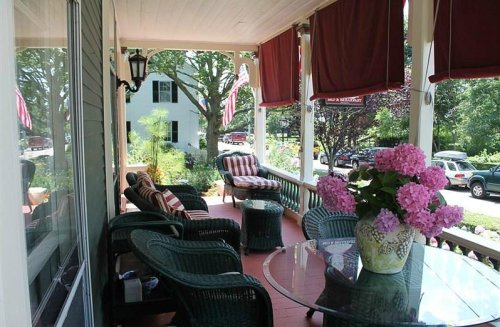 A cozy porch with wicker furniture, a glass table, and pink hydrangeas, surrounded by greenery and parked cars.