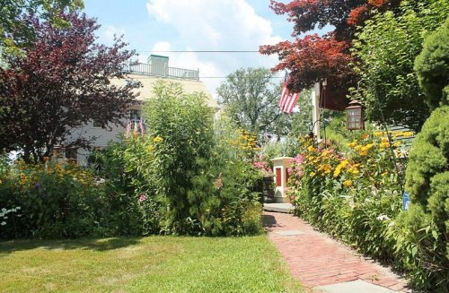 A vibrant garden path lined with colorful flowers, leading to a house with an American flag and lush greenery.