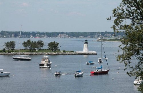 A scenic view of a harbor with boats and a lighthouse on a small island, surrounded by calm waters and trees.