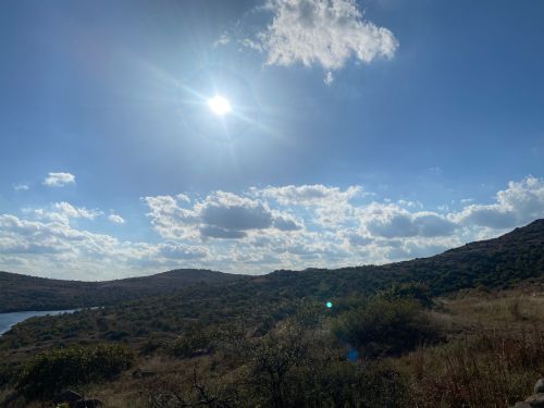 A bright sun shines over a hilly landscape with scattered clouds and a serene body of water in the distance.