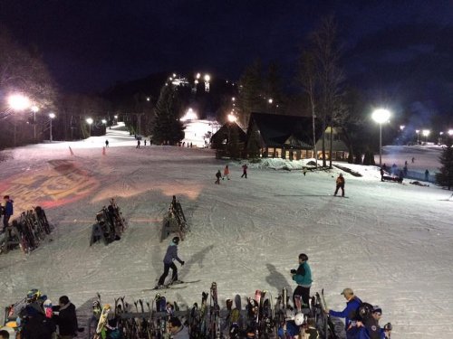 Skiers on a snowy slope at night, with a lodge and illuminated ski trails in the background.