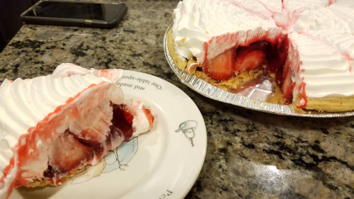 A slice of strawberry pie on a plate next to the whole pie, featuring whipped cream and a glossy red filling.