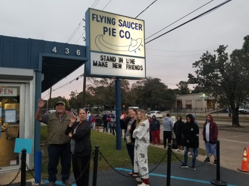 A group of people waves in front of the Flying Saucer Pie Co. sign, with a line of customers waiting outside.