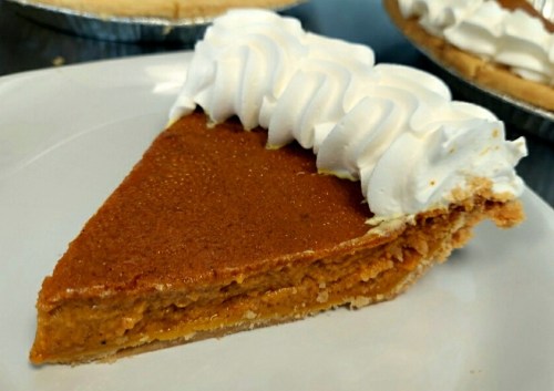 A slice of pumpkin pie topped with whipped cream on a white plate, with more pies in the background.