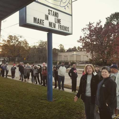 A long line of people waiting outside, with a sign above reading "Stand in New Lines." Trees and buildings in the background.