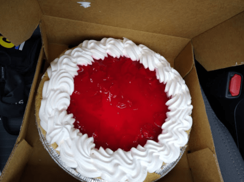 A cherry pie with a glossy red topping and whipped cream edges, placed inside a cardboard box.