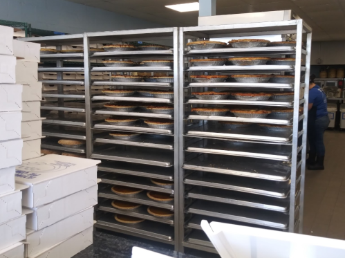 Racks filled with baked pies in a bakery, with boxes stacked nearby and a person working in the background.