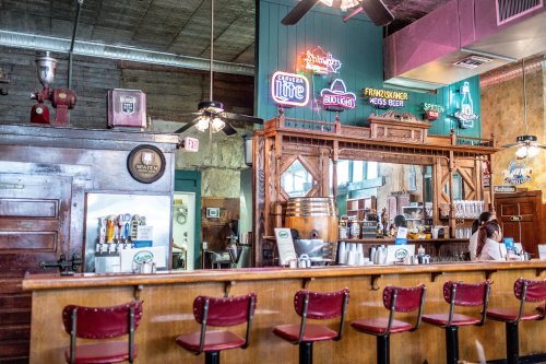 A rustic bar with wooden stools, neon signs, and a vintage decor, featuring a beer tap and a cozy atmosphere.