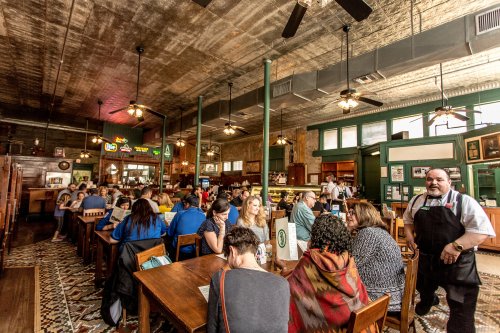 A bustling restaurant interior with diners at wooden tables and a waiter in an apron, featuring vintage decor and ceiling fans.