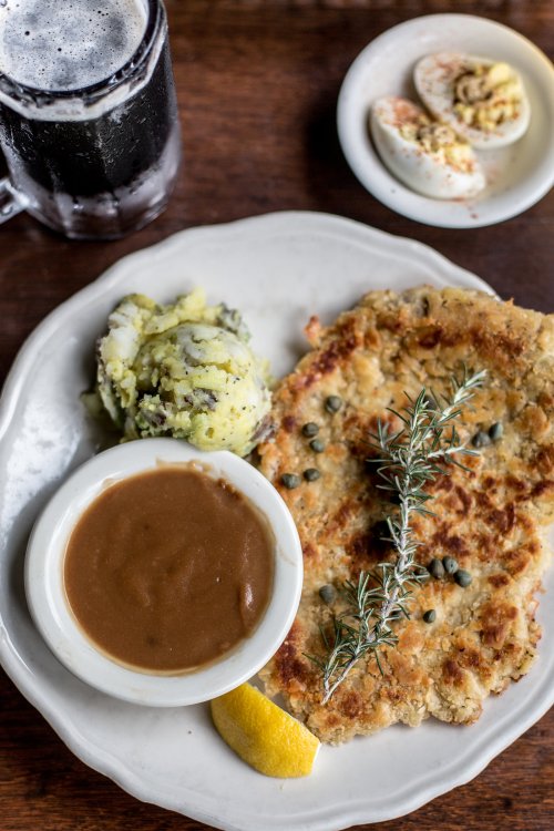 A plate featuring a breaded fish fillet, mashed potatoes, brown gravy, a lemon wedge, and a sprig of rosemary.