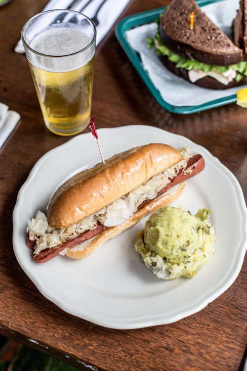 A hot dog topped with sauerkraut on a plate, accompanied by a side of potato salad and a glass of beer.
