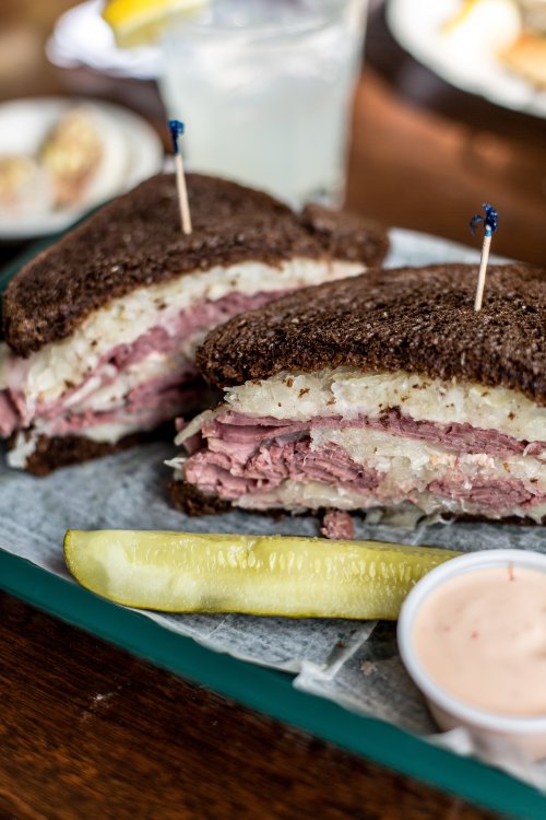A close-up of a sliced sandwich with layers of meat and sauerkraut on dark bread, served with a pickle and dipping sauce.
