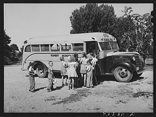 A vintage school bus with children gathered around it in a rural setting.