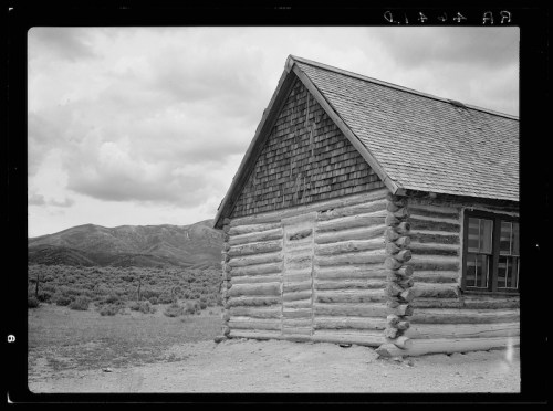 A rustic log cabin with a shingled roof, set against a backdrop of mountains and cloudy skies.