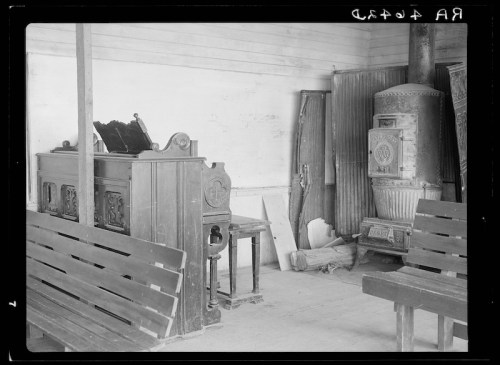 Interior of a rustic room featuring a wooden piano, a stove, and wooden benches against a plain wall.