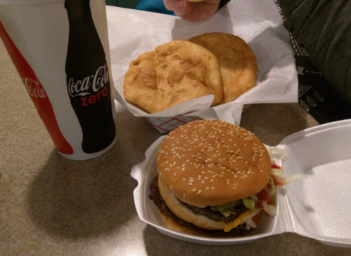 A sesame seed burger with toppings, two fried dough pieces, and a large cup of Coca-Cola Zero on a table.