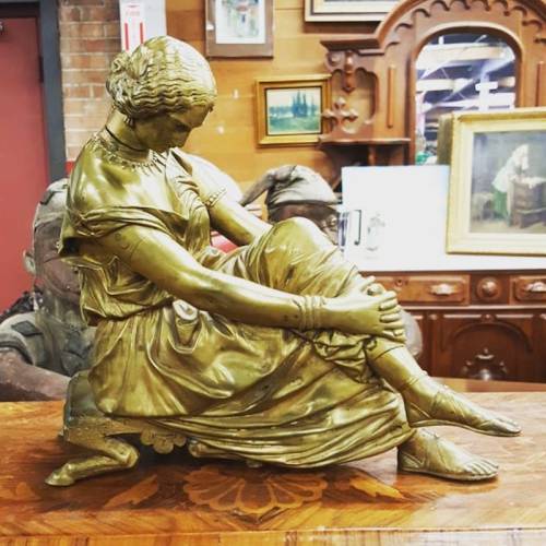 A bronze sculpture of a seated woman in a flowing dress, resting her arm on her knee, displayed on a wooden table.