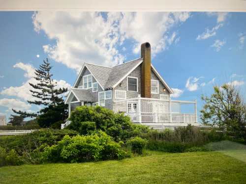 A charming gray house with a tall chimney, surrounded by greenery and under a blue sky with fluffy clouds.