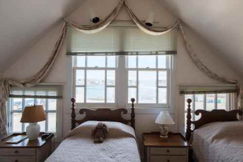 Cozy attic bedroom with two twin beds, lamps, and a teddy bear, featuring large windows with scenic views.