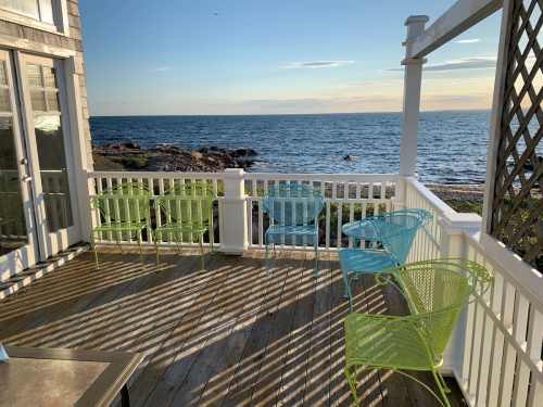 A seaside deck with colorful chairs overlooking the ocean at sunset, casting long shadows on the wooden floor.