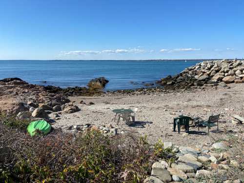 A serene beach scene with rocks, a calm sea, and a few chairs set up on the sand under a clear blue sky.
