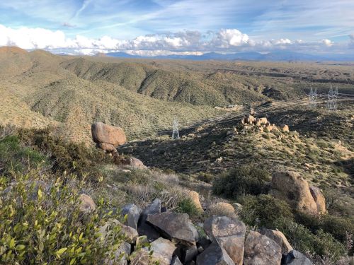 A panoramic view of rolling hills and rocky terrain under a blue sky with scattered clouds. Power lines stretch across the landscape.