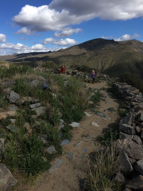 Two people walk along a rocky path surrounded by greenery and mountains under a partly cloudy sky.