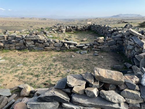 Stone walls form a circular structure on a rocky landscape, with distant hills and a clear sky in the background.