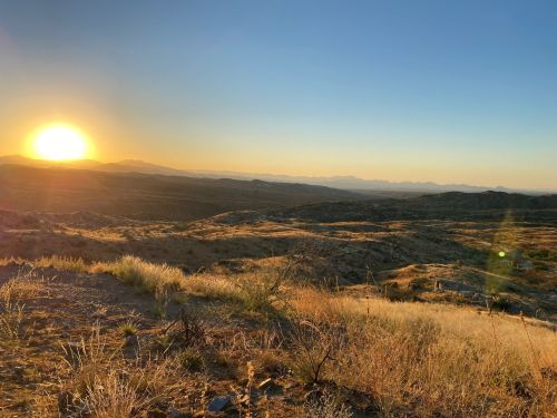 A sunset over a vast desert landscape, with rolling hills and sparse vegetation under a clear sky.