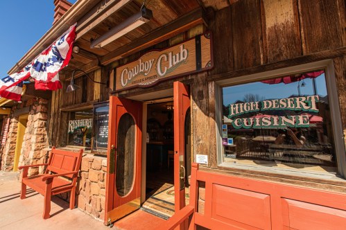 Exterior of the Cowboy Club restaurant, featuring wooden walls, red doors, and a sign for "High Desert Cuisine."