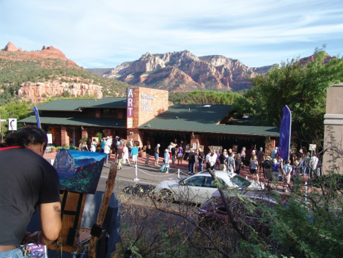 A crowd gathers outside an art gallery in a scenic area with mountains in the background. An artist paints nearby.