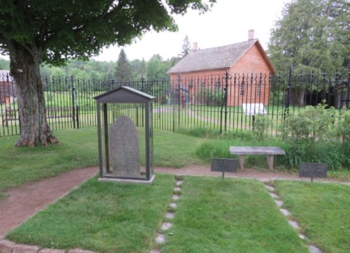 A historic grave marker surrounded by grass, with a wooden building and fence in the background.