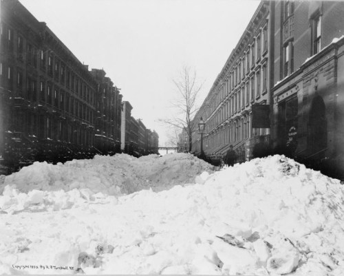 A historic black-and-white photo of a snow-covered street, with large snowdrifts between tall buildings.