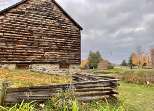 A rustic wooden barn with a stone foundation, surrounded by greenery and autumn foliage under a cloudy sky.