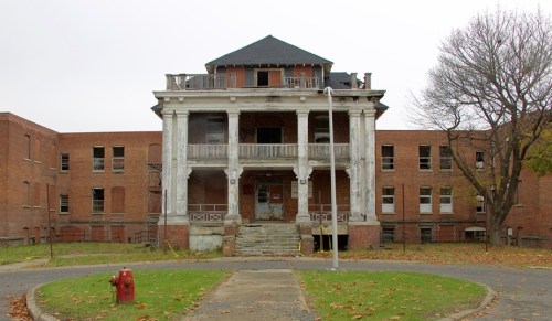 Abandoned building with a grand facade, featuring columns and a dark roof, surrounded by overgrown grass and trees.