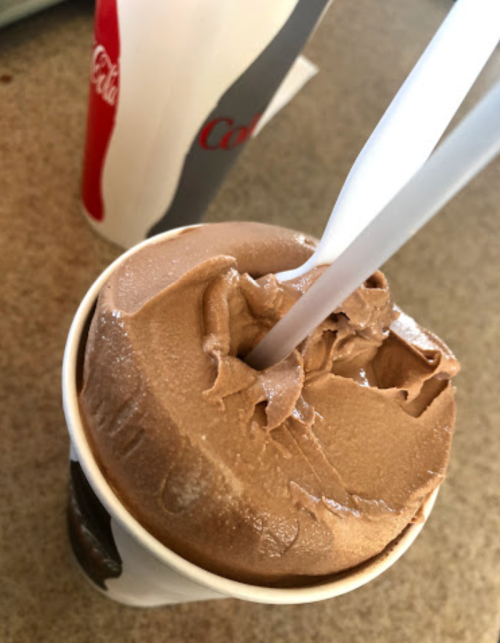 A cup of chocolate soft serve ice cream with two plastic spoons, next to a fast food drink cup.