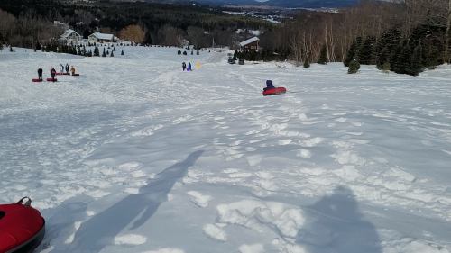 A snowy hill with people tubing down, surrounded by trees and distant houses under a clear blue sky.