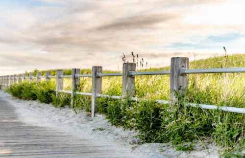 A wooden fence along a sandy path, surrounded by tall grass and a cloudy sky.