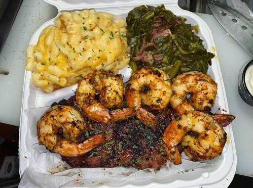 A takeout container with grilled shrimp, macaroni and cheese, collard greens, and pulled pork.