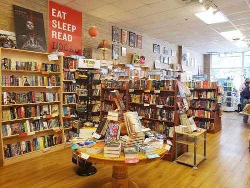 A cozy bookstore with wooden shelves filled with books, a central table stacked with titles, and a "Eat Sleep Read Local" sign.