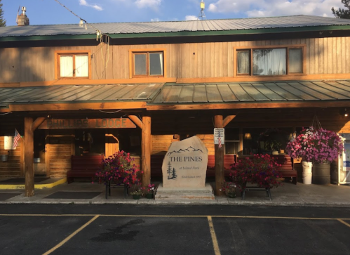 A rustic lodge with a wooden exterior, flower pots, and a stone sign reading "The Pines" in a scenic setting.