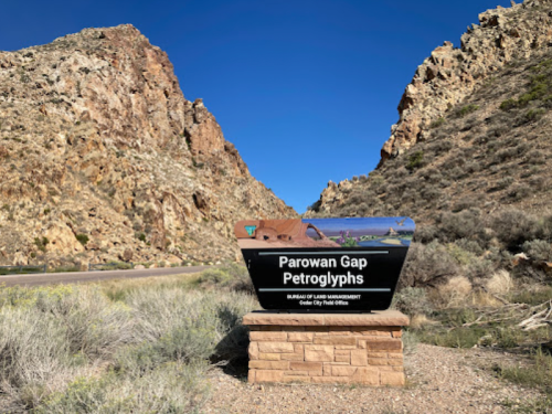 Sign for Parowan Gap Petroglyphs, surrounded by rocky cliffs and desert vegetation under a clear blue sky.