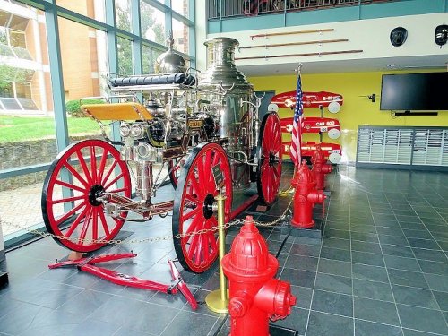 A vintage fire engine displayed indoors, surrounded by fire hydrants and a U.S. flag, with large windows in the background.