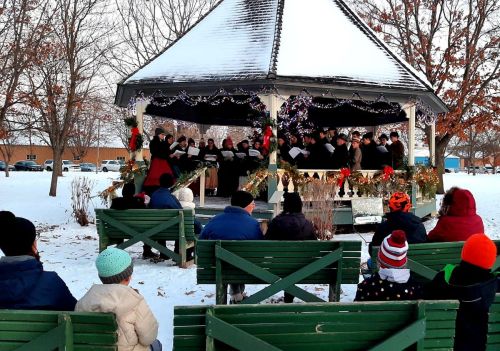 A winter scene with a choir performing in a gazebo, while an audience sits on benches in the snow.