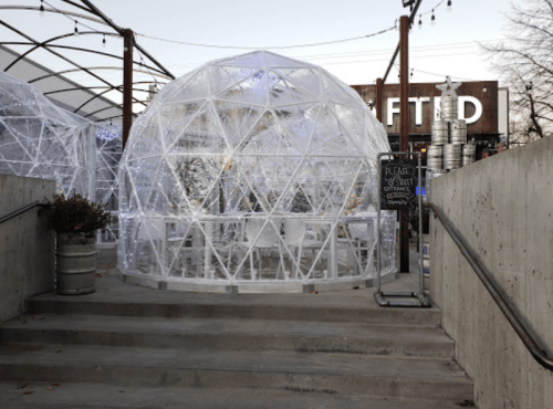 A clear geodesic dome structure on a patio, surrounded by string lights and a rustic building in the background.